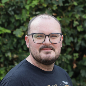 Head and shoulders portrait of James with short hair, glasses and facial hair, wearing a dark T-shirt, with a blurred green foliage background